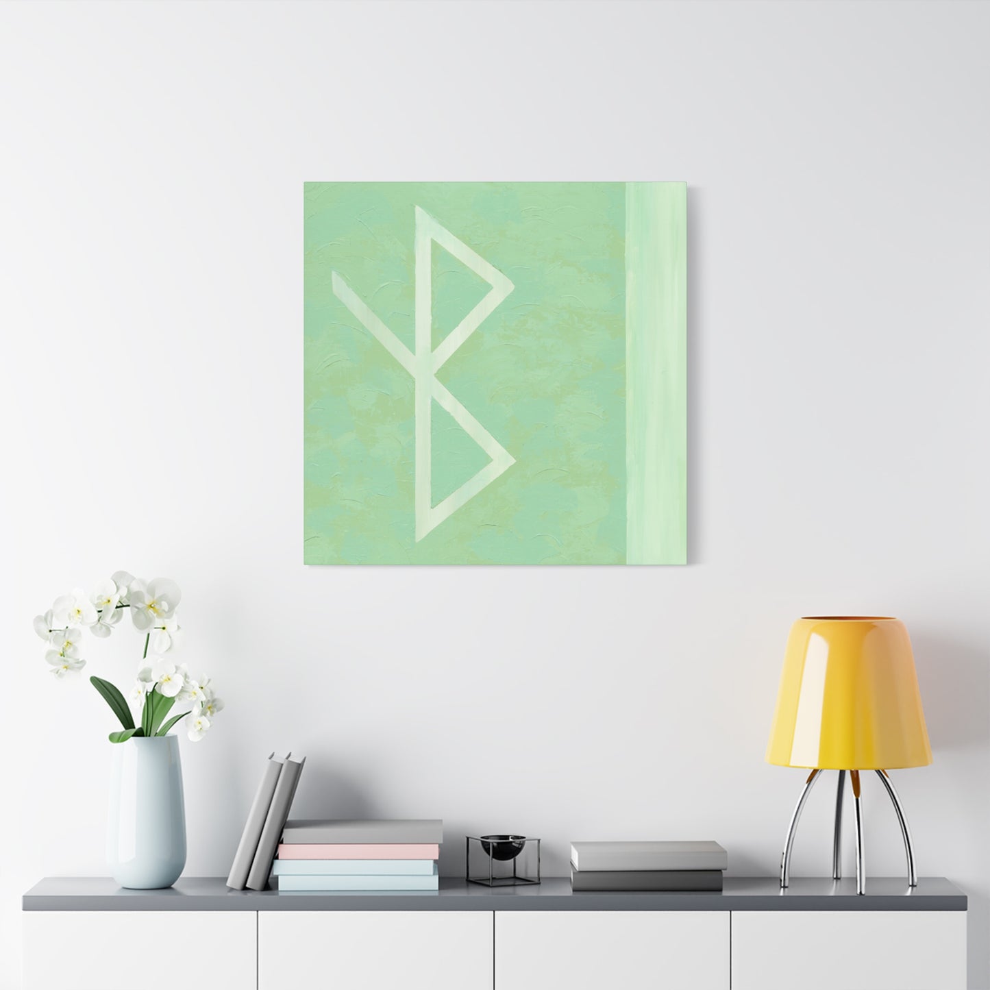 A square abstract wall art print with a mint green background and a large white bind rune symbol for health, displayed above a console table with a yellow lamp, books, and a vase of white flowers.