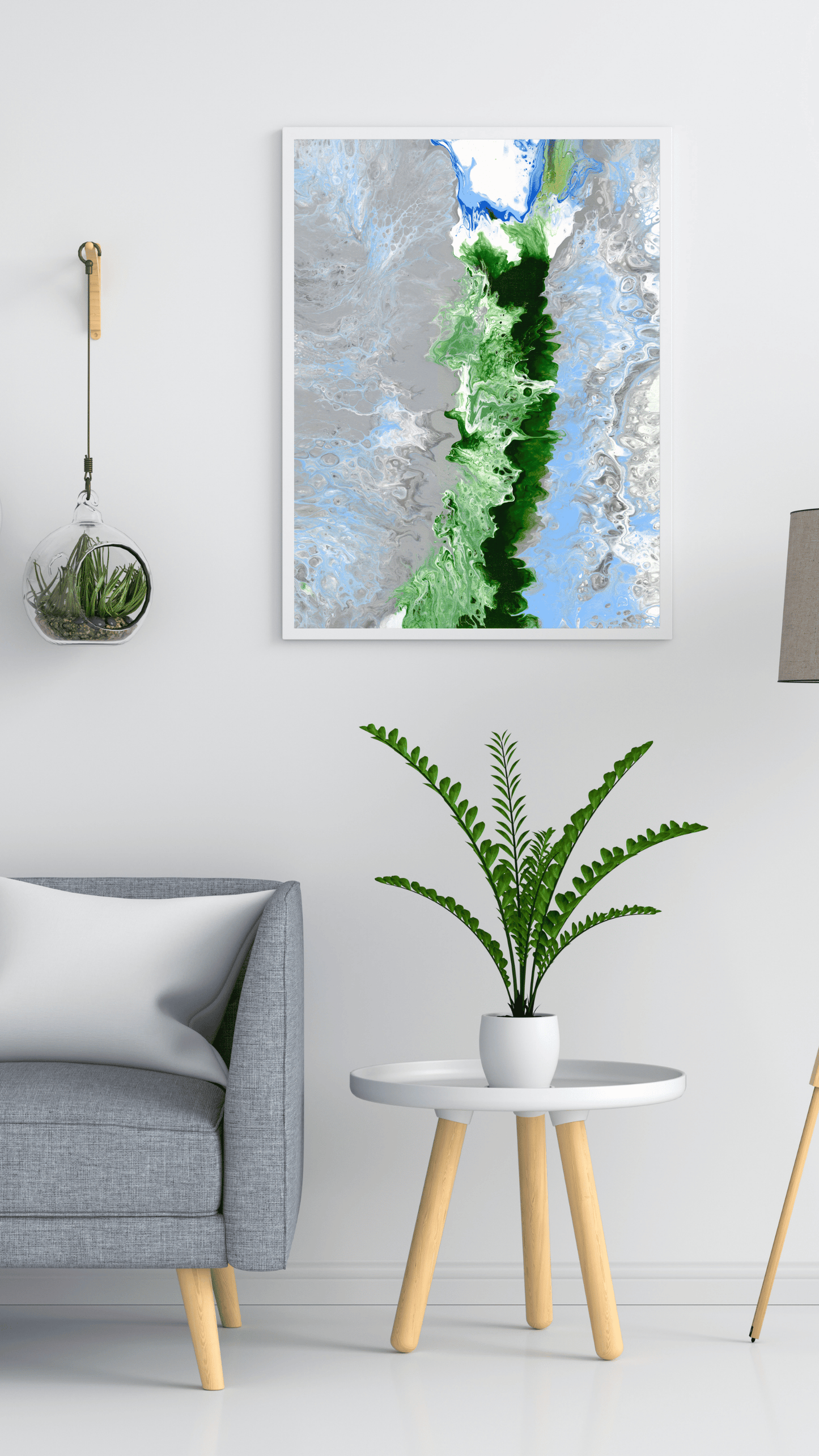 A framed abstract art print featuring a vertical green and blue textured design, displayed on a white wall in a modern living room setting above a gray armchair and a small side table with a potted fern.