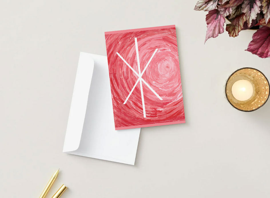 A greeting card with a red and pink abstract swirl design and white geometric lines of a Bind Rune that symbolizes Strength, shown with a white envelope on a light beige background with decorative props including dried leaves, a candle, and gold pens.